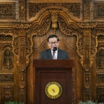 "Pejabat Pemprov DKI Jakarta berbicara di podium dalam acara resmi, dengan latar belakang ukiran kayu tradisional Indonesia di Kedutaan Besar Republik Indonesia di Seoul."